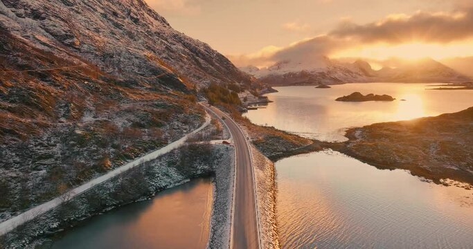 Aerial view of road, sea and snowy mountains at sunset in winter. Lofoten Islands, Norway. Top drone view of beautiful bridge, reflection in water, rocks, orange sky with clouds and golden sunlight