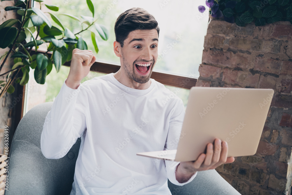 Poster photo of impressed lucky young guy wear white shirt rising fist winning game modern gadget indoors a