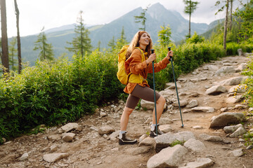 Young female traveler with hiking poles and a bright backpack enjoys the mountain scenery along a...