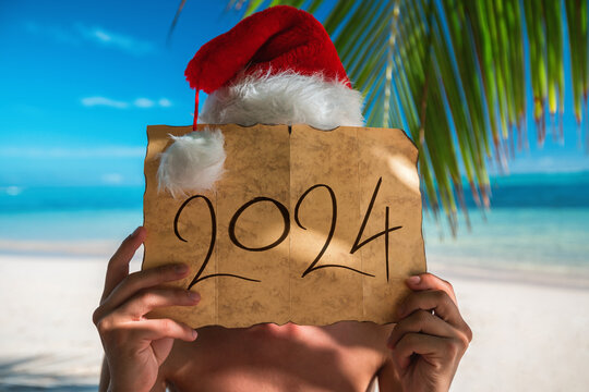 Winter seasonal vacation on a tropical island for the new year 2024, man with Santa Claus hat and treasure old map on exotic beach with palm trees.