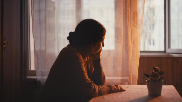 Sad grandmother sitting at table and looking out the window at sunset, nostalgia