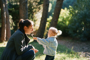 Mom blows on dandelions in the hand of a little girl squatting in the forest