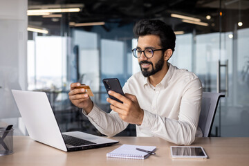 Young smiling Indian businessman sitting in office at desk and using credit card and mobile phone