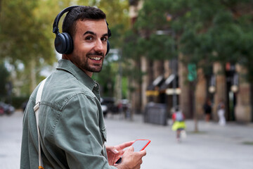 Cheerful portrait of a caucasian young man walking outside in the street, using phone to listen music with headphones, turns back to look at the camera. Copy space. - obrazy, fototapety, plakaty