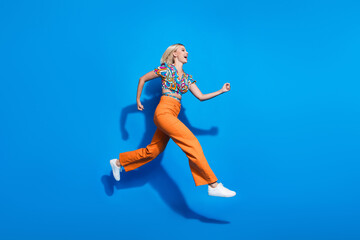 Full length photo of positive cheerful girl dressed print top orange pants run shopping at empty space isolated on blue color background