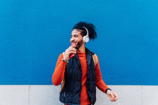 Pensive hispanic man using mobile and headphones standing looking away in a blue urban wall