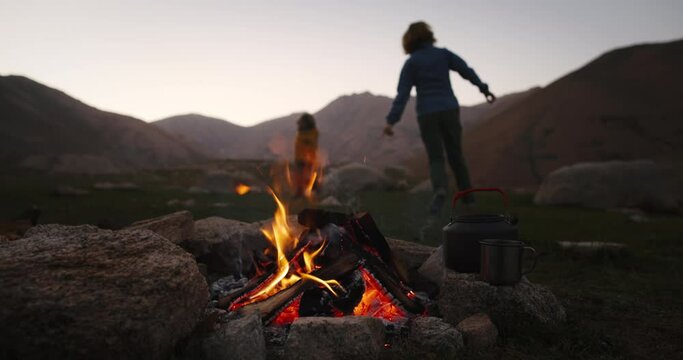 Child and woman playing catch-up by fire. Mom and son playing together in background, bonfire burning in foreground. Happy cheerful funny woman and child campers hikers playing catch-up together.