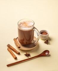 Glass mug of hot chocolate with whipped cream and cinnamon stick on a yellow background with anise star, spoon and cocoa powder.