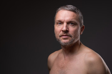 .Slightly smiling face of a middle-aged man with a beard looking at the camera. Close-up portrait of a fifty-year-old man with a naked torso against a dark background.