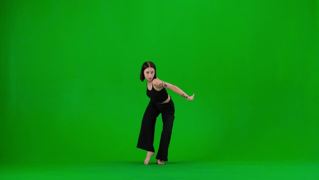 In the frame on a green background, a limp. Dances young beautiful girl. Demonstrates dance moves in the style of hip hop. She stares at the camera. She is feminine, barefoot in a black top and pants