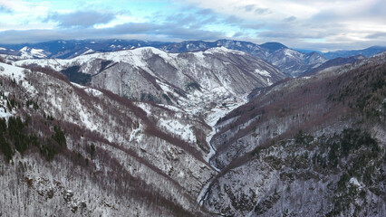Aerial panorama above a valley inside the mountains covered with snow. The beech woodlands are frozen and the mountain peaks at the horizon are covered in snow as it is winter. Carpathia, Romania.