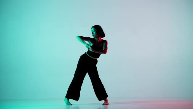 In the frame on a blue, light background. Dances young, beautiful girl. Demonstrates dance moves in the style of hip hop. Shes staring at the camera. Shes feminine in a black top and pants