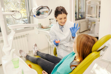 A happy dentist is holding jaw model and talking to a girl in chair at dentist office.