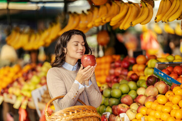 A woman is standing at farmers market and smelling fresh organic apple.