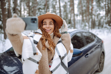 Beautiful woman stands next to a car against the backdrop of a snowy forest with a phone in her hands. Young tourist takes a selfie with her phone next to her car on a snowy road. Travel concept.