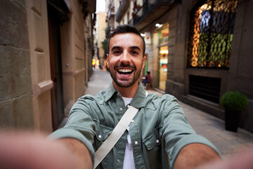 Happy selfie of a young caucasian man in an old town of Barcelona. Male tourist taking a self portrait using smartphone to post it on social media, - obrazy, fototapety, plakaty