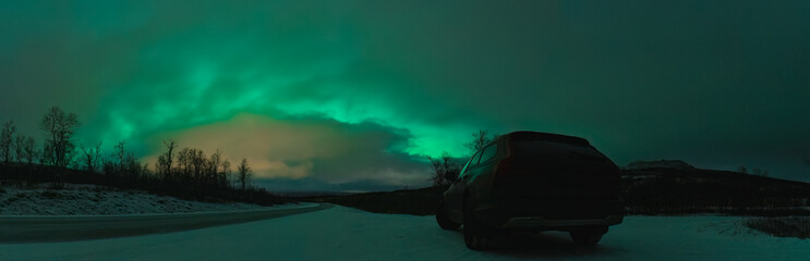 Scenic frosty night panorama of dark car on mountain road in front of Northern green lights shine...