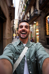 Happy vertical selfie of a young caucasian man in an old town of Barcelona. Male tourist taking a self portrait using smartphone to post it on social media, - obrazy, fototapety, plakaty