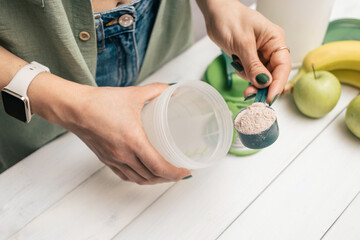 Young woman in jeans and shirt with measuring spoon in her hand puts portion of whey protein powder into shaker on white wooden table with bananas and apples. Process of making protein drink