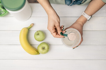 Young woman with measuring spoon in her hand puts portion of whey protein powder into shaker on...