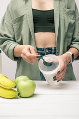 Young woman in jeans and shirt with measuring spoon in her hand puts portion of whey protein powder...