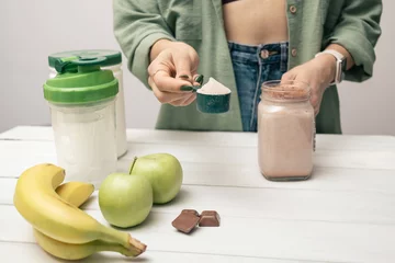  Young woman in jeans and shirt holding measuring spoon with protein powder, glass jar of protein drink cocktail, milkshake or smoothie above white wooden table with chocolate pieces, bananas, apples © O.Farion
