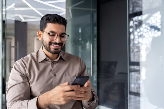 Businessman holding phone inside office, joyful man smiling uses smartphone app at workplace, browses social networks, and writes text message