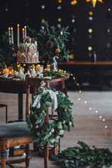 Christmas elegant composition with handmade wreath on wooden chain and beautiful homemade gingerbread house, candles on the table. Family party festive decoration, cozy atmosphere, aesthetics