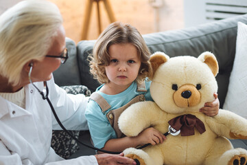 Kind senior female paediatrician doctor examining little child, cute girl during home visit or clinic check up. Concept of healthcare, medical assistance, insurance. Prescribing treatment for disease