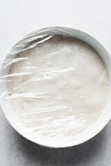 Focaccia dough in a white bowl covered with plastic wrap, process of making focaccia, dough rising...