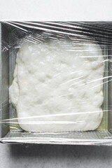 Focaccia dough in a square baking tin covered with plastic wrap, process of making focaccia, dough rising in a baking pan