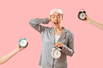Shocked young woman in pajamas and female hands with alarm clocks on pink background