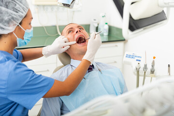 Female dentist in face mask inspects male patient teeth with mirror and probe