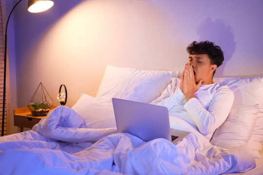 Sleepy young man using laptop in bed at night