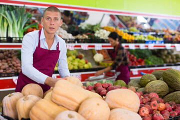 European man in uniform standing among shelves in greengrocer putting pumpking on counter.