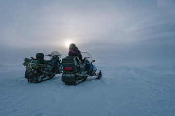 Scenic view at two snowmobiles stand close at snowy and windy mountain top. One person in overall...