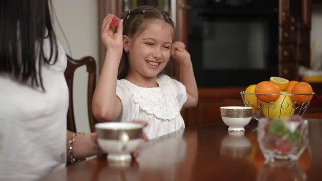 Sweet joy of a child. A happy girl, sitting at the table with her mother, takes a candy and shows it to the camera