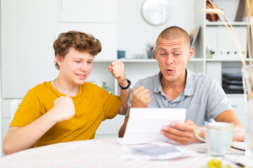 Son and his father sitting at the table and reading letter that contains great news.