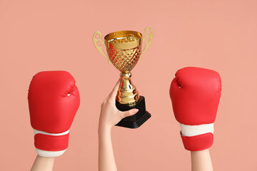 Female hands with gold cup and boxing gloves on pink background