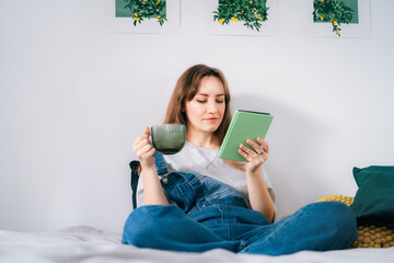 Young woman reading e-book on tablet reader in bed. Female relaxing with hot drink while reading an...