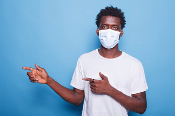 Afro haired black man in portrait facing the camera and gesturing with his hands to the right. African American guy with a face mask marketing a product while motioning with his arms.