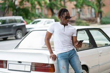 stylish young african american male standing near his retro car and using smartphone