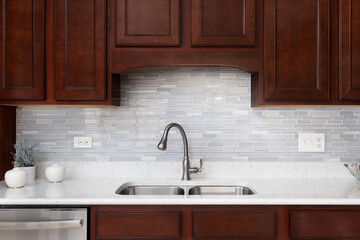 A kitchen faucet detail with a glass tiled backsplash, dark wood cabinets, and a white marble...