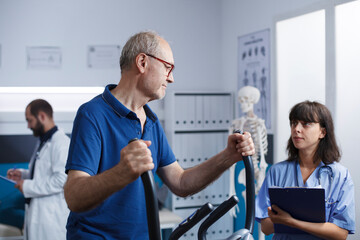 Obraz premium Medical nurse helps elderly patient who is receiving therapy with alternative medicine while exercising on stationary bike. Health professional with clipboard examines old man for rehabilitation