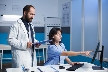 Caucasian doctor and female nurse in blue scrubs and lab coat collaborate in clinic office, using...