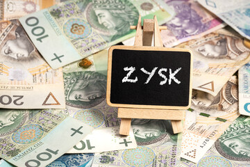 a small wooden writing board standing on scattered Polish zloty PLN banknotes, a chalk inscription "Zysk" on the black board