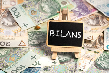 a small wooden writing board standing on scattered Polish zloty PLN banknotes, a chalk inscription "Bilans" on the black board.