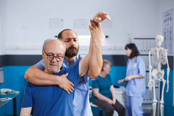 Medical nurse stretching muscles and treating physical injuries by elevating arm of elderly person. At physiotherapy consultation, male osteopath does mobility exercises with senior patient.