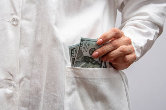 A woman doctor puts several hundred dollar bills in her pocket and a white doctor's coat.  Concept: illegal enrichment, bribe to a health worker, bribery of officials.
