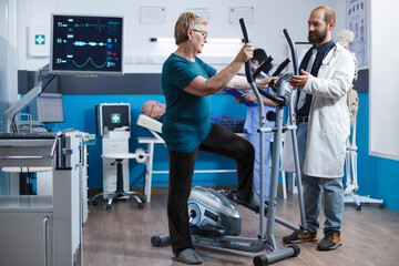 Elderly woman uses electric bicycle for physical rehabilitation after being hurt. Senior female patient using a stationary bike for gymnastics while the doctor assists with physiotherapy.